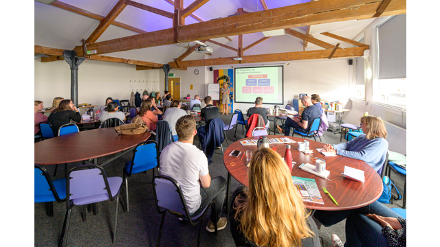 Groups of people sat around tables with a presenter in front of a screen