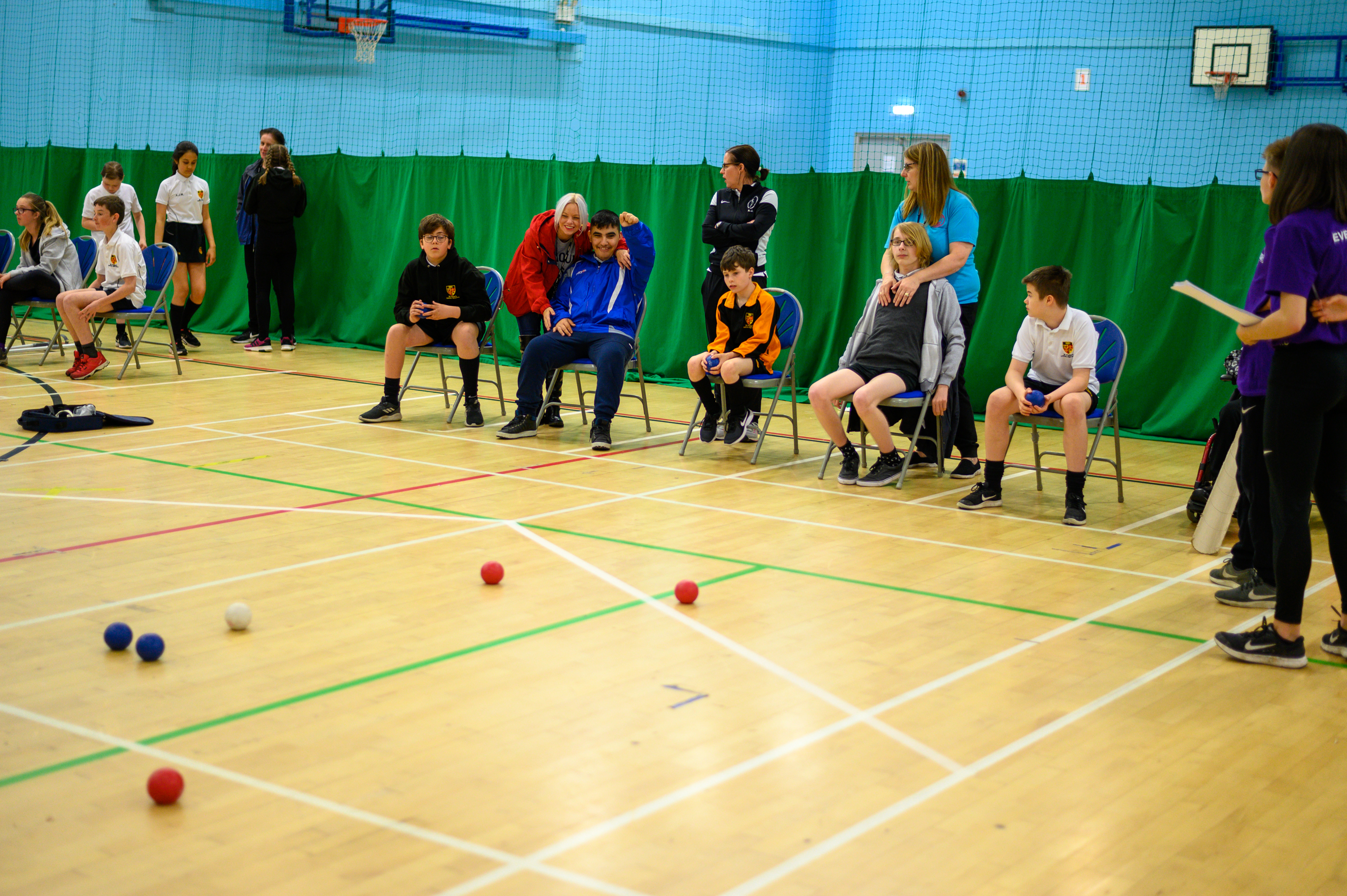 Children sat in chairs in a sports hall playing boccia