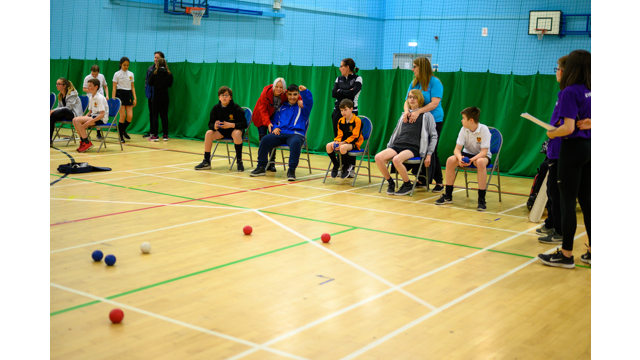 Children sat in chairs in a sports hall playing boccia