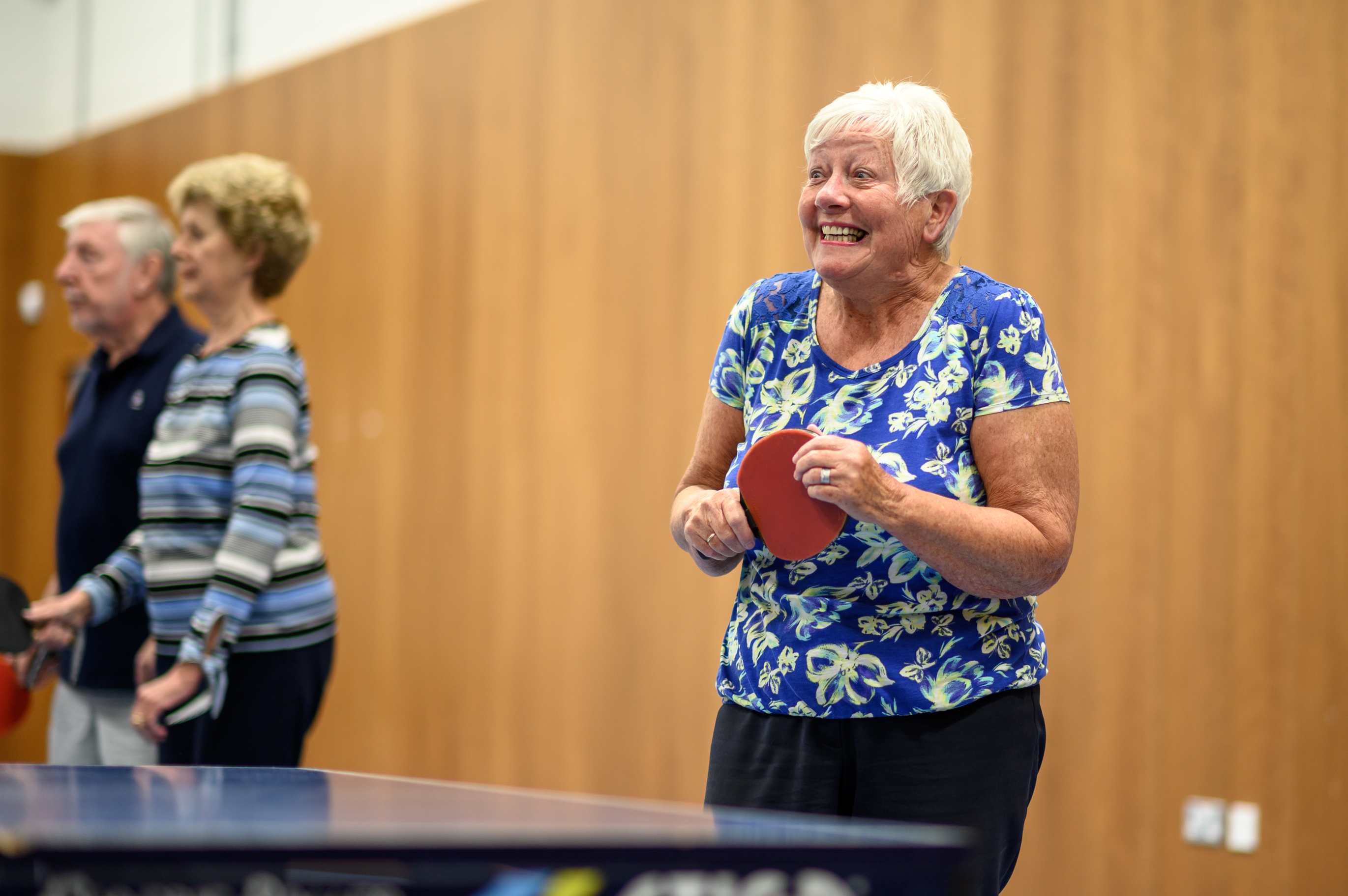 Older adult stood smiling holding a table tennis bat at a table