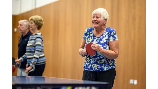Older adult stood smiling holding a table tennis bat at a table