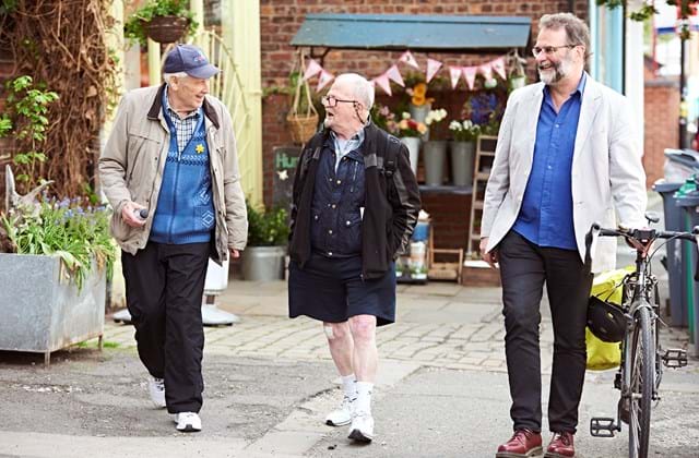 Three older adults walking and talking along a pavement, one is pushing a bike.