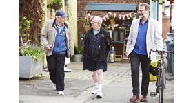 Three older adults walking and talking along a pavement, one is pushing a bike.