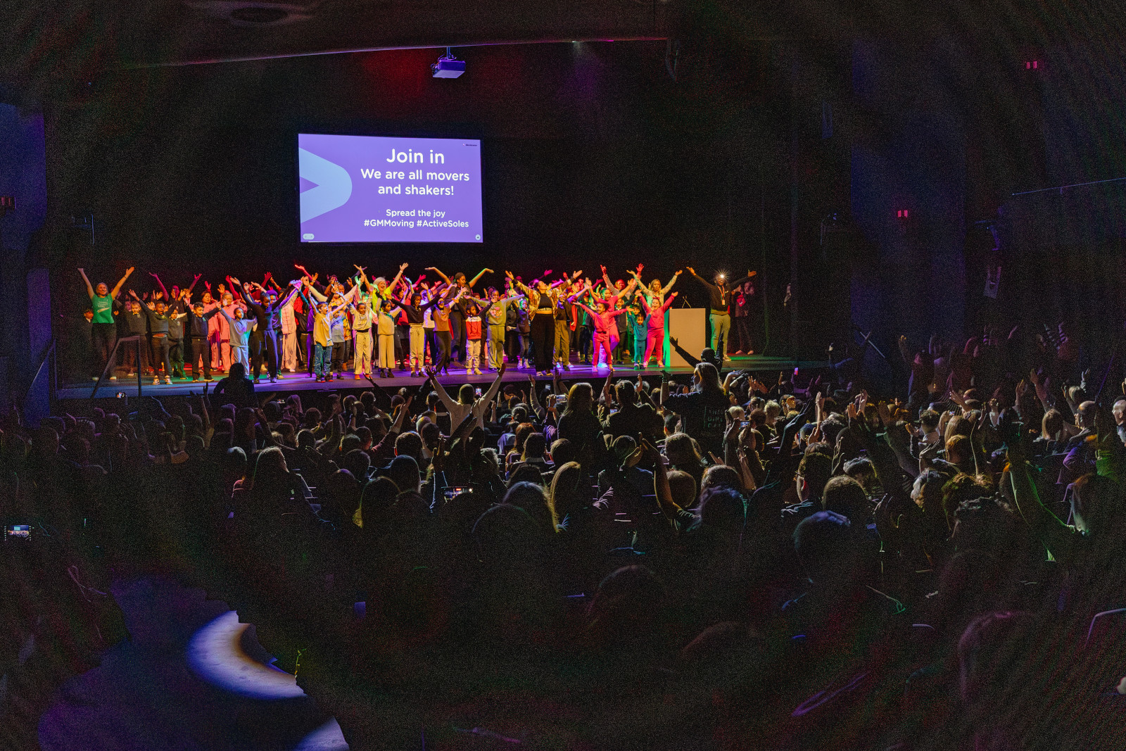 A crowd of young people dressed in bright colours raise their hands in unison on a conference stage at a theatre.