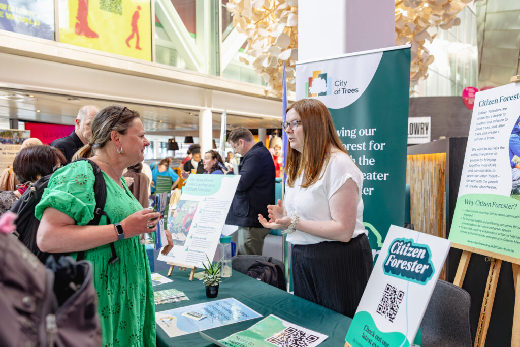 Two people talk on opposite sides of a conference booth for 'Citizen Forester', which has leaflets and full size banners about the project and City of Trees.