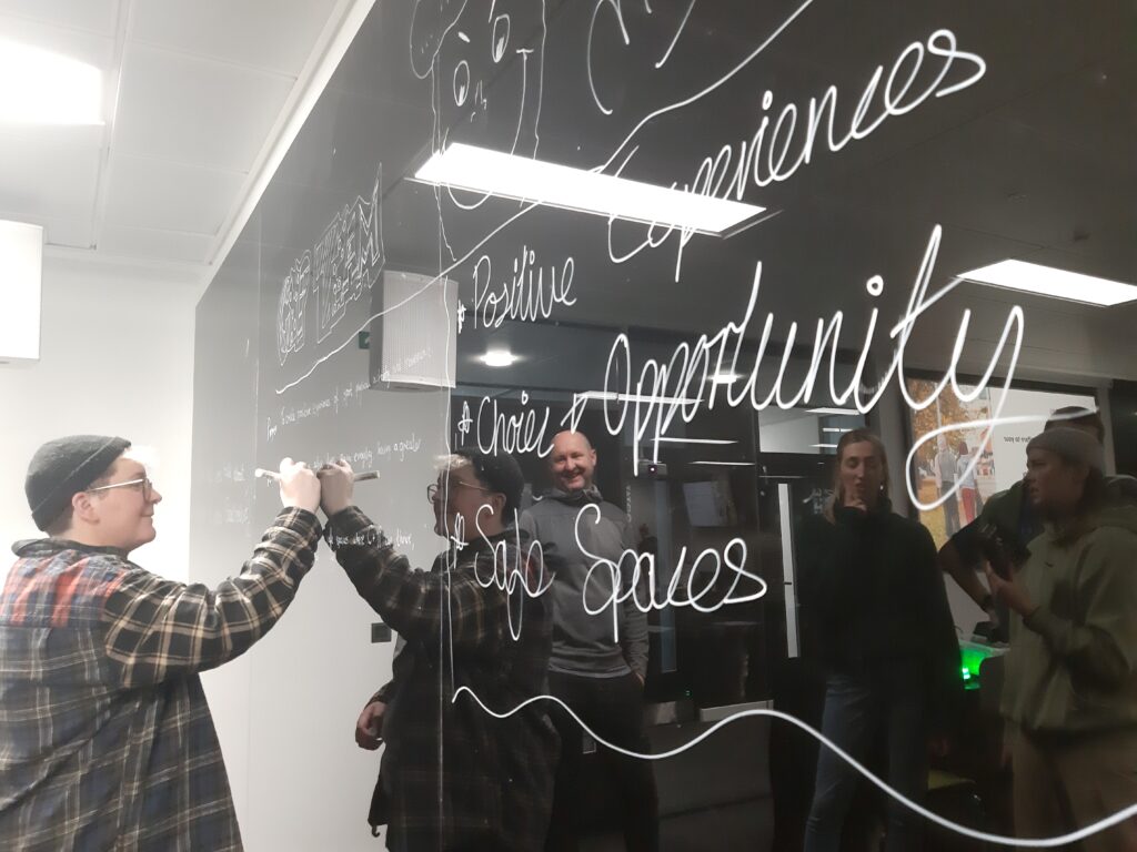 Jess Eastoe writes on a glossy blackboard, with the words 'Safe Spaces' and 'Opportunity' visible.