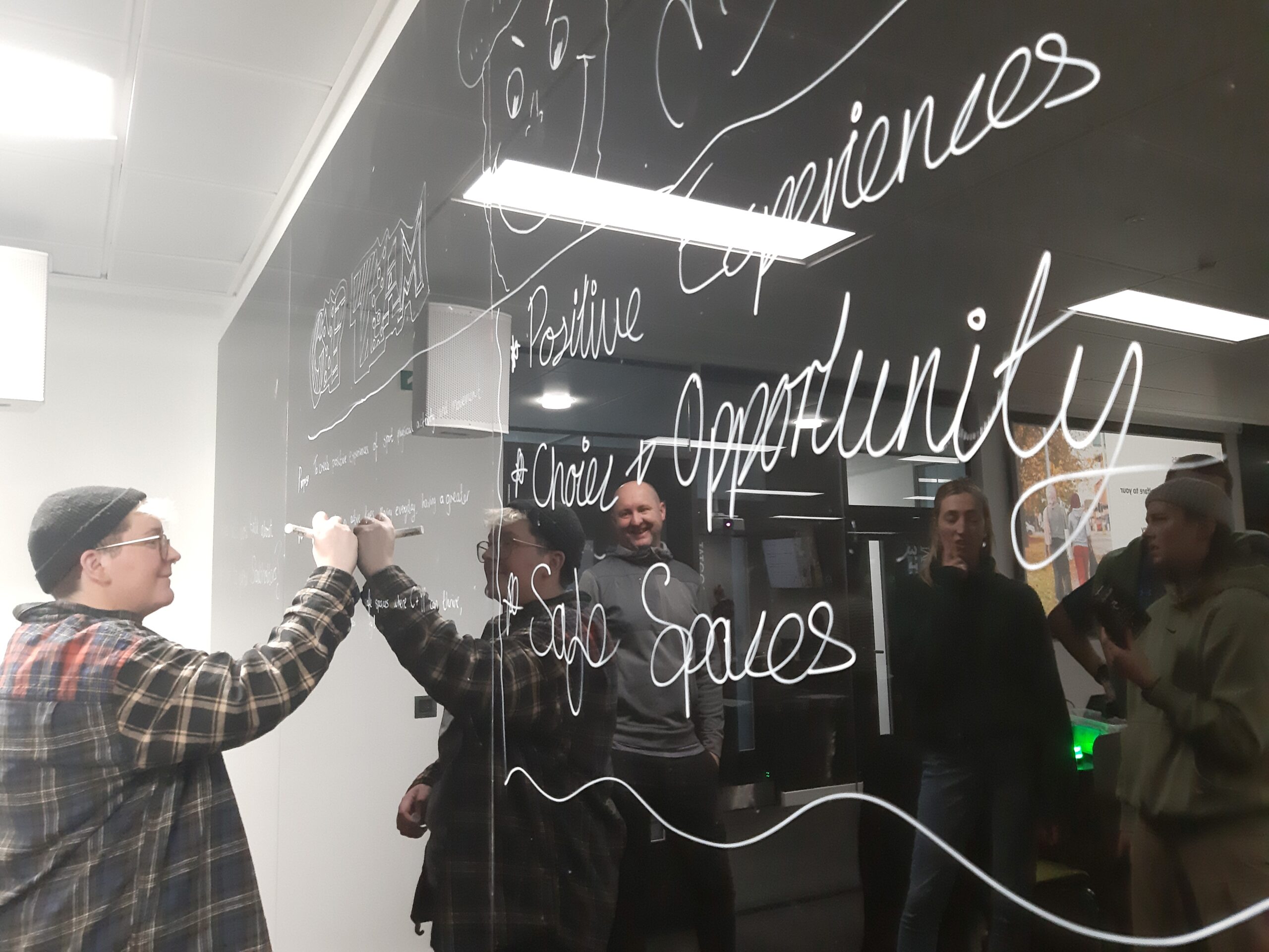 Jess Eastoe writes on a glossy blackboard, with the words 'Safe Spaces' and 'Opportunity' visible.