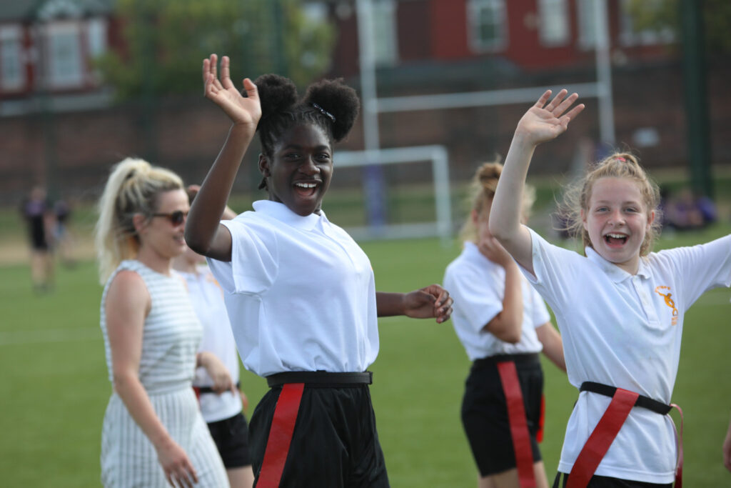 Two schoolgirls smiling and waving to the camera outside during an activity.