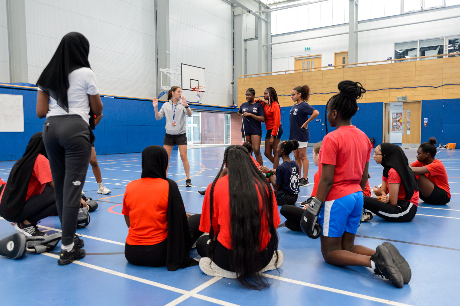 A group of young people stand, sit and kneel in a semi circle when listening to a trainer in a sports hall.