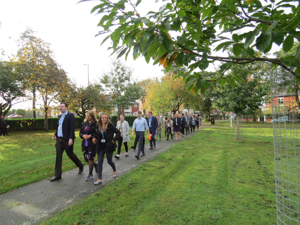 A large group of office workers walk along a path in a leafy park on a sunny day.
