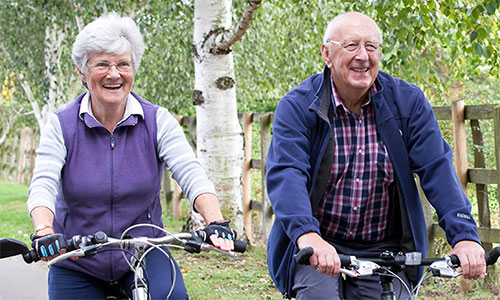 An older couple cycle through a green and leafy park