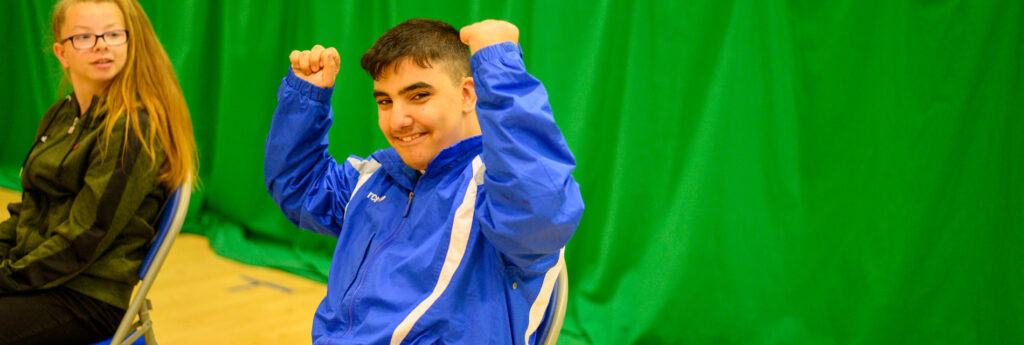 A school child sits in a school hall, raising his fists in celebration