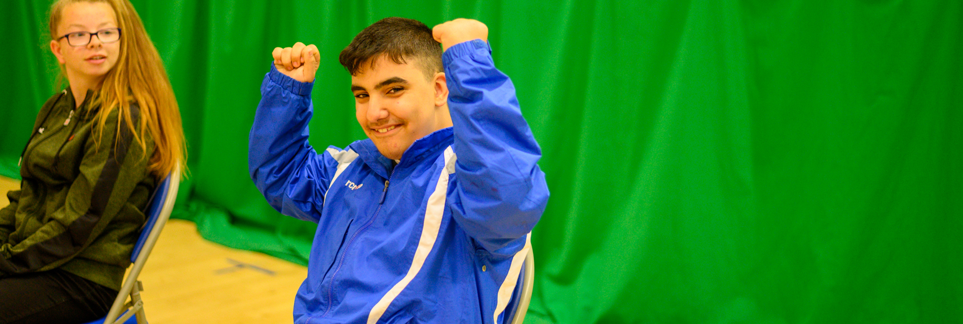 A school child sits in a school hall, raising his fists in celebration
