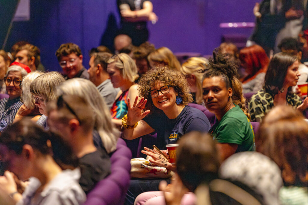 A person waving towards the camera in crowded theatre seating at a conference