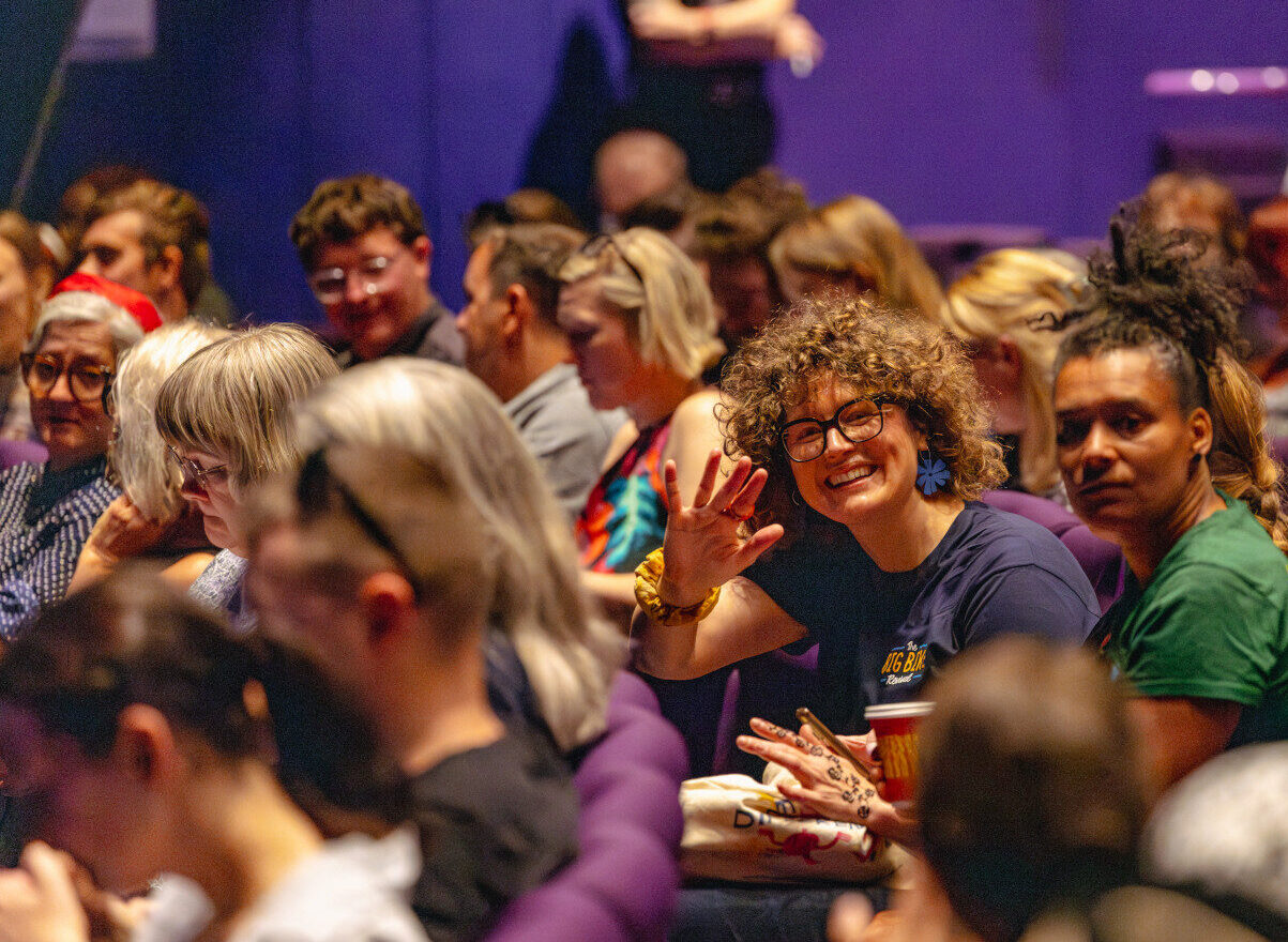 A person waving towards the camera in crowded theatre seating at a conference
