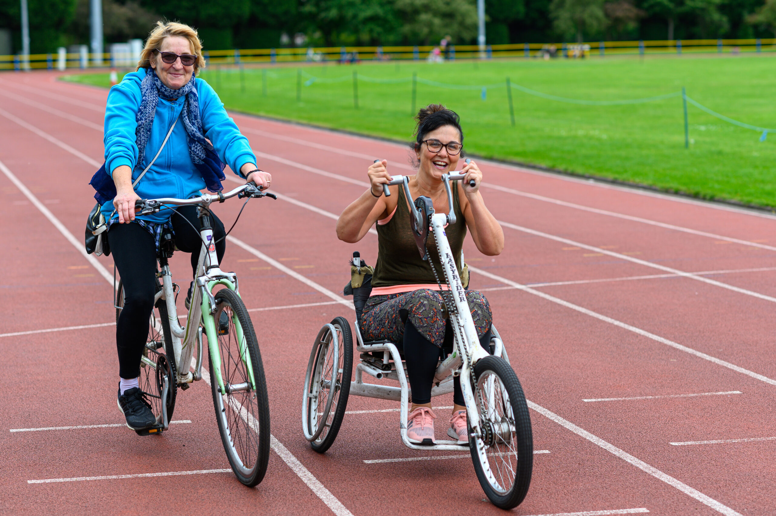 Two adults cycle round an outdoor track during an inclusive cycling session with adaptive bikes.