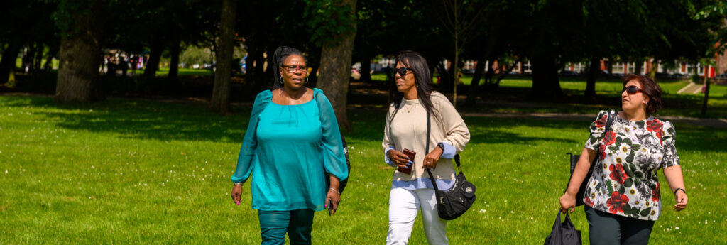 Three women walk on a sunny day through a green and leafy park