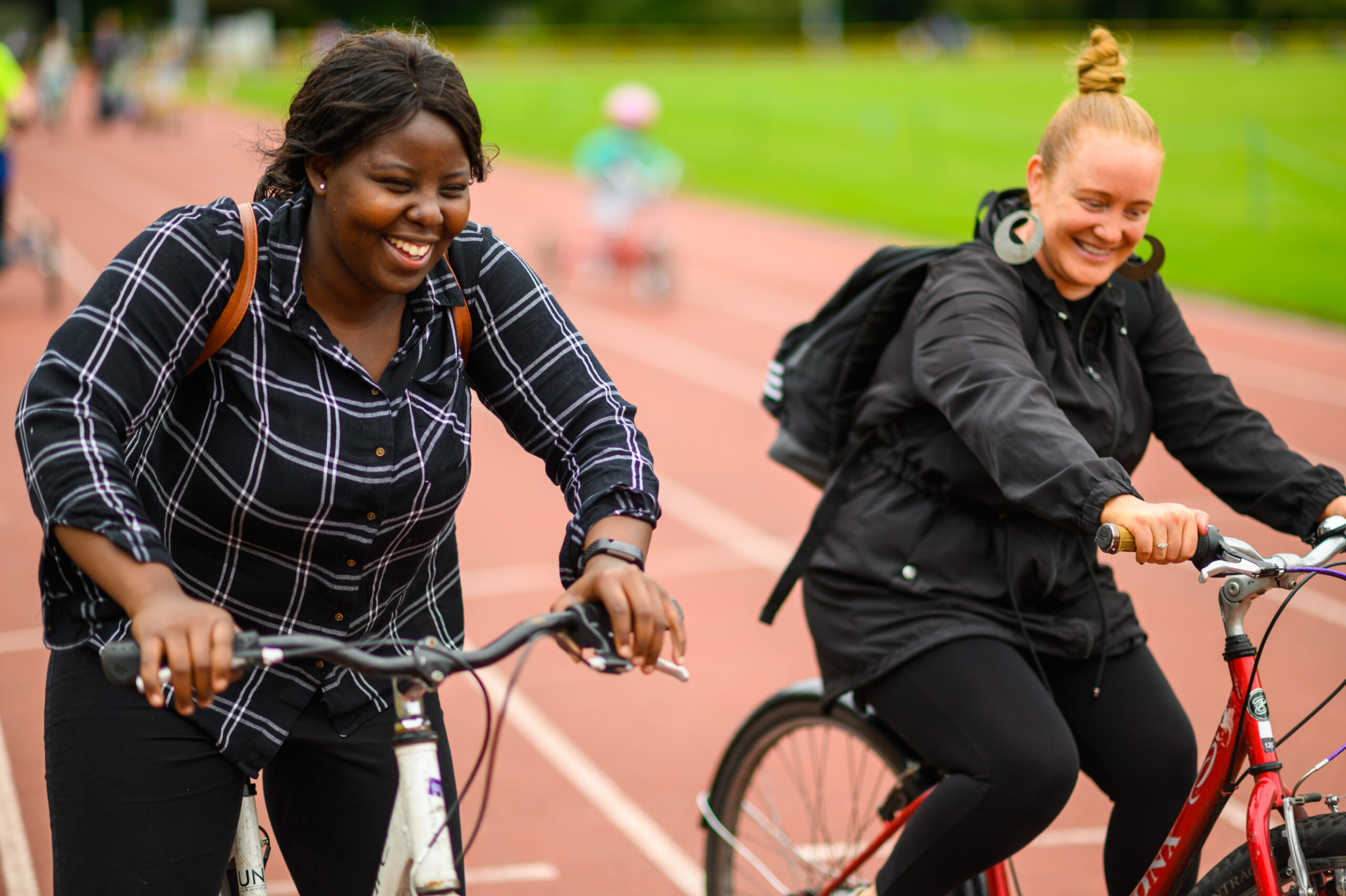 Two young women smile and ride bikes on an outdoor sports track