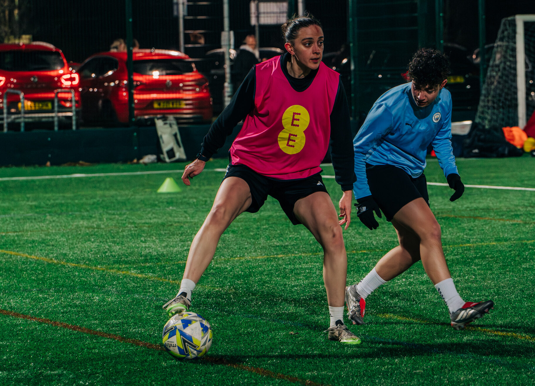 A girl in a pink bib controls a football with an opponent in blue close by.