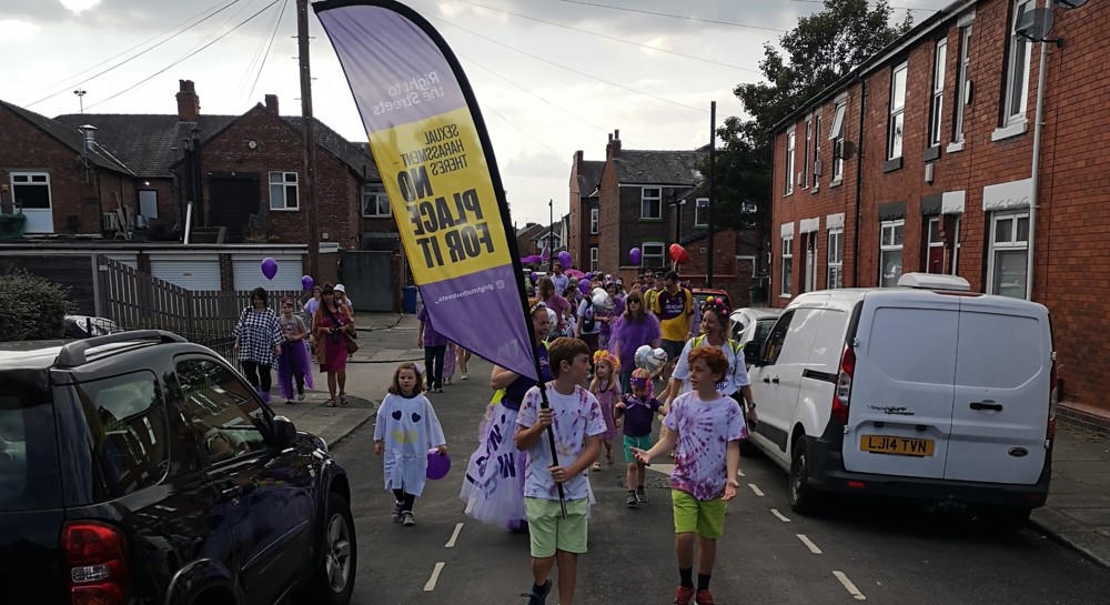 Young women and girls march through a street, holding a sign that says 'No Place For It'.