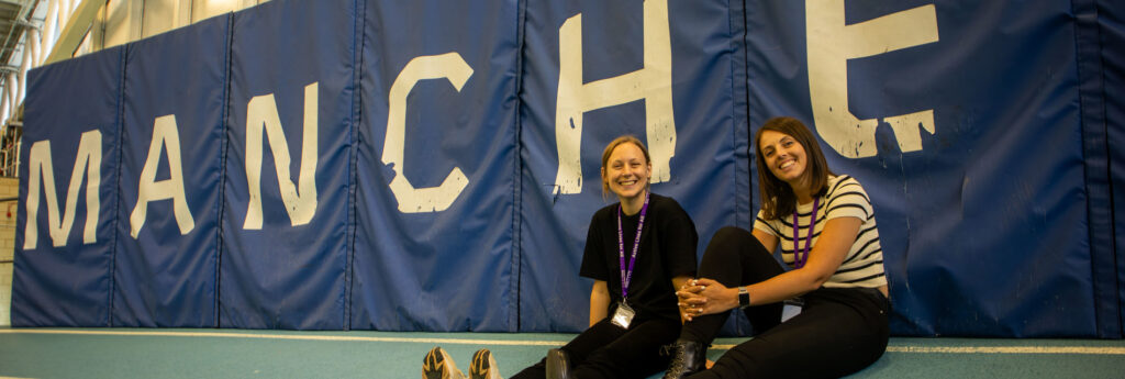 Two sports welfare leads sit in front of a sign that reads, 'Manchester'.