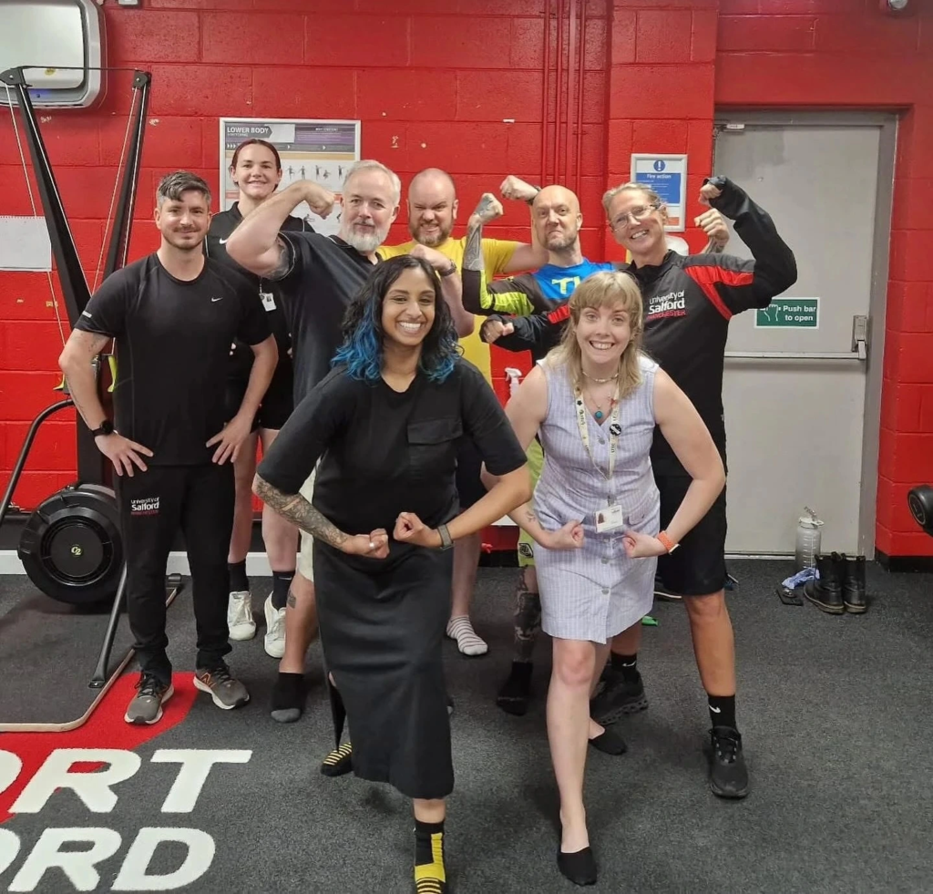 A diverse group of adults in a gym laugh and stand in body-builder poses