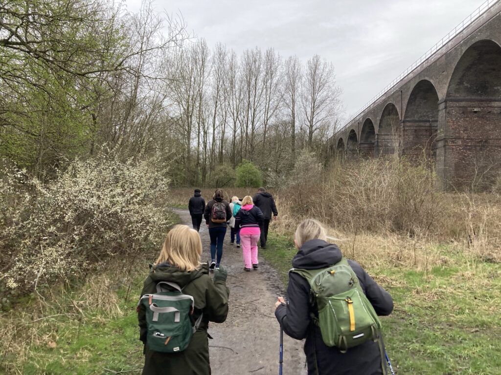 A group of adults walk through a green park near an old Victorian bridge