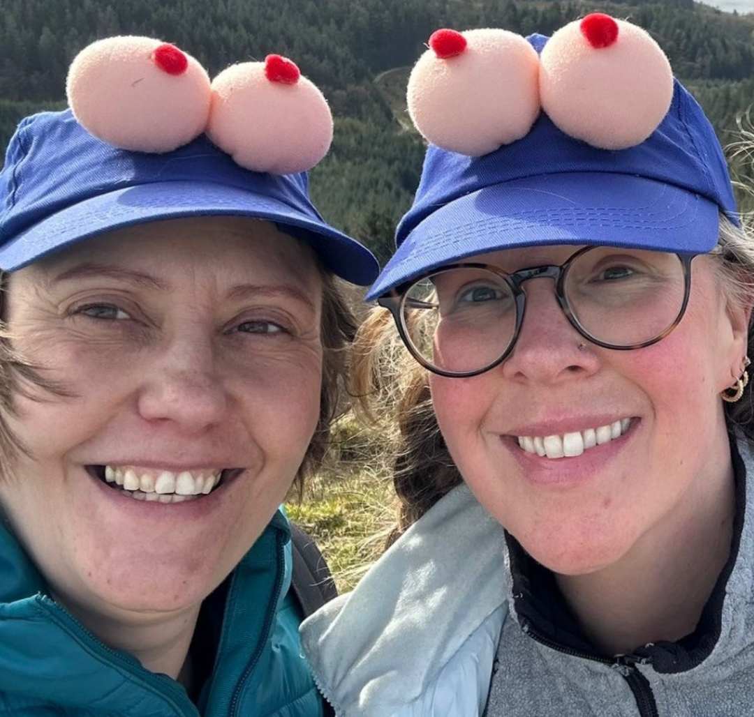 Two women smile while wearing caps for breast-cancer awareness while hiking in the Peak District