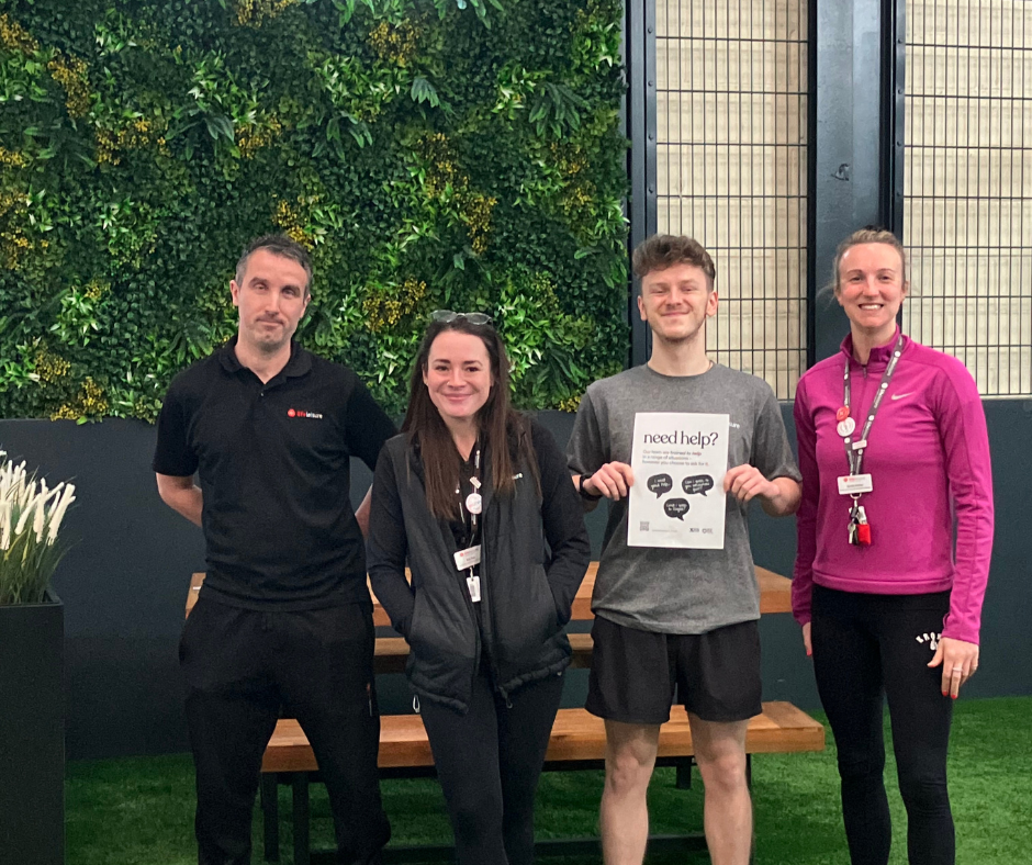 Four adults stand and smile in athletic wear. One person is holding a sign that reads, 'Need help?'. Alistair Dixon, Eve Fox, Shaun Adamson, Sarah Potter. Staff members at Life Leisure Houldsworth Village and Support Office.