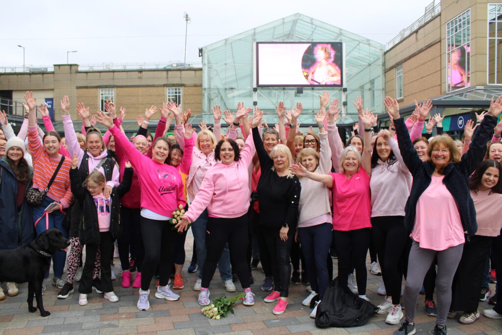 A group of women in Stockport wear pink t-shirts and wave at the camera. They are joined by instructors Sue Mellor and Laura Latham with the rest of the flashmob participants in the centre of Merseyway.