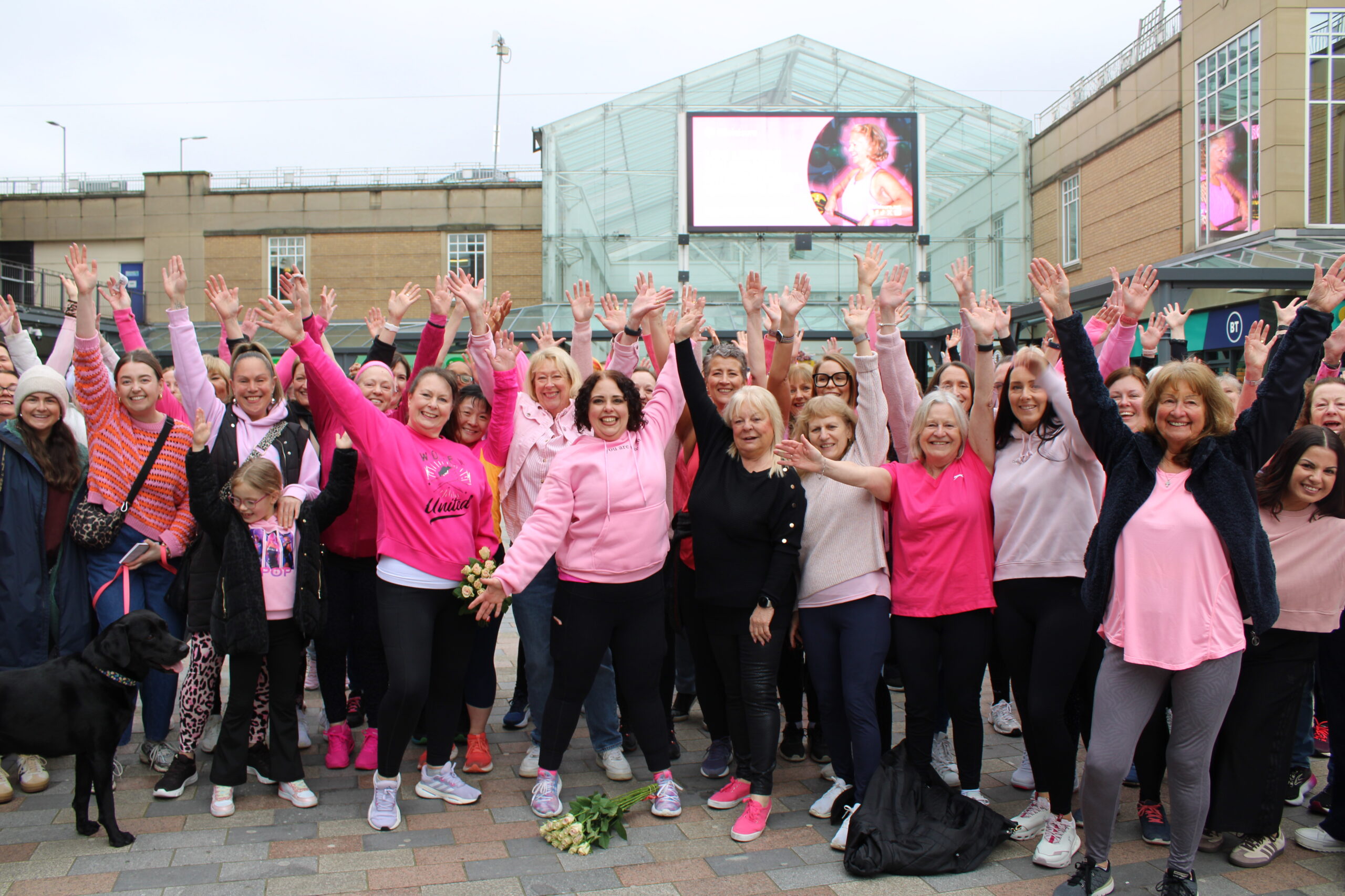 A group of women in Stockport wear pink t-shirts and wave at the camera. They are joined by instructors Sue Mellor and Laura Latham with the rest of the flashmob participants in the centre of Merseyway.