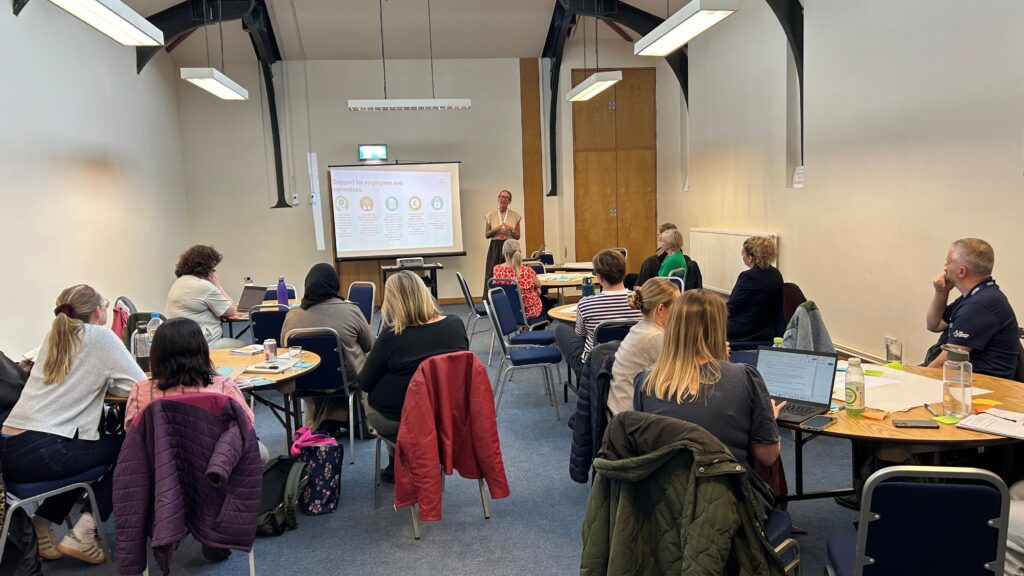 A photograph of the third GM Moving and Noisy Cricket social care event with a speaker using a slide show and audience sat around tables.