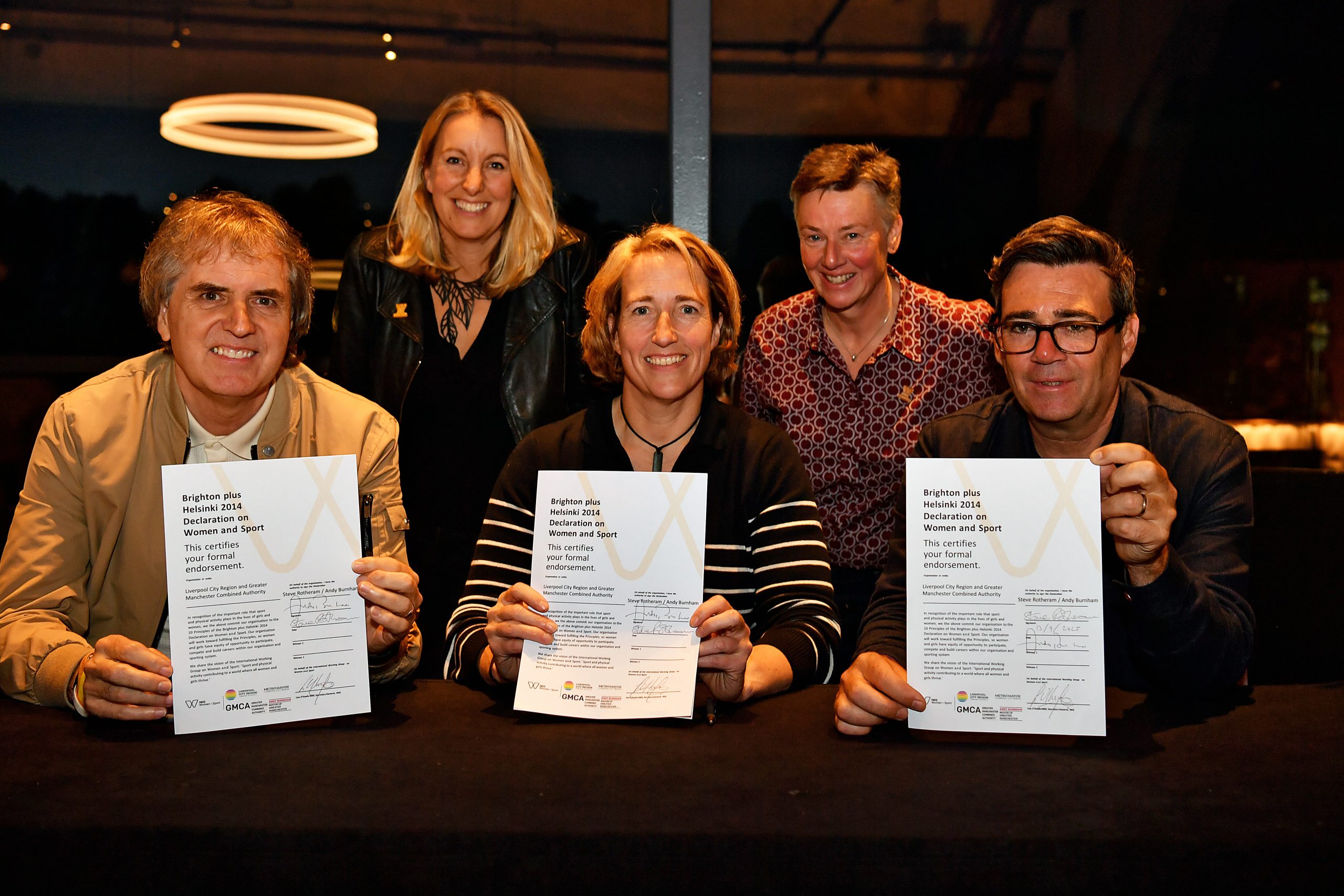 Liverpool City Region Mayor Steve Rotheram and Greater Manchester Mayor Andy Burnham sit at a table holding signed copies of the Brighton Plus Helsinki Declaration alongside representatives from the IWG on Women and Sport