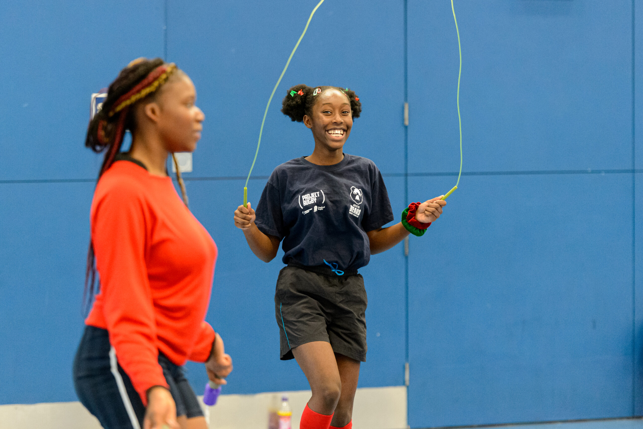 A girl in a sports hall uses a jump rope while smiling at the camera