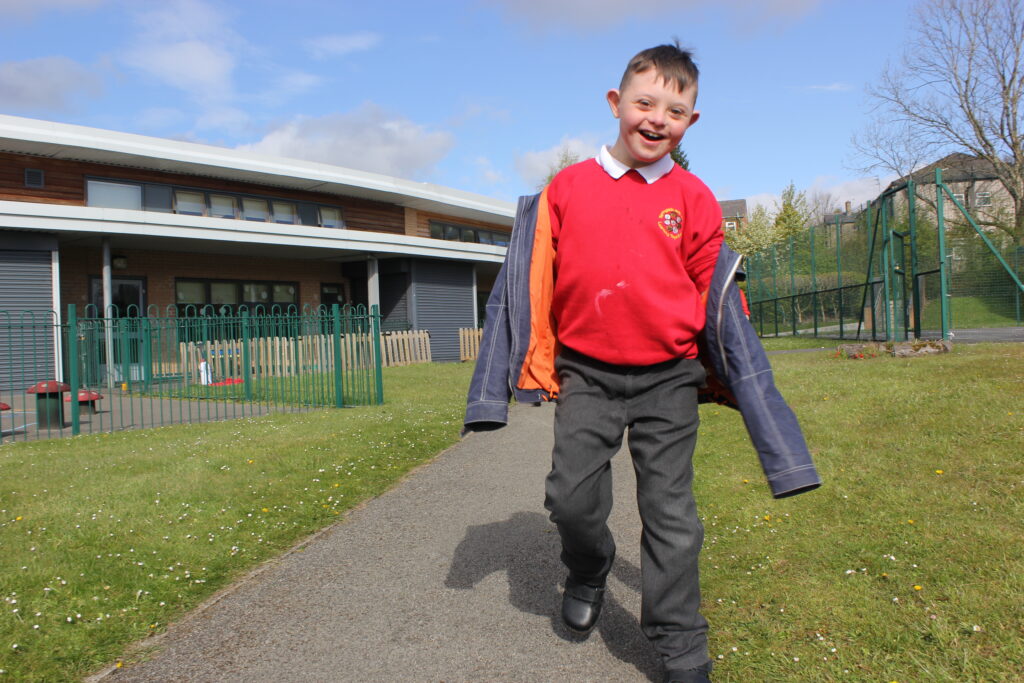 A smiling boy in school uniform walks towards the camera on a sunny day