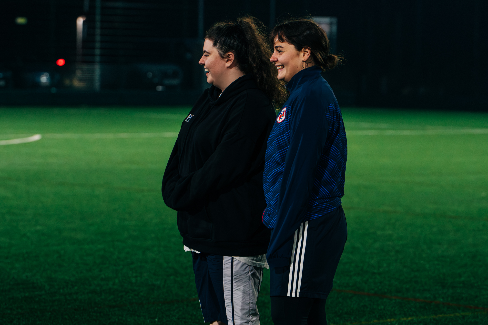 Two people smile while watching a sports match on an outdoor pitch at night time